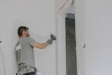 Gri üniformalı ve eldivenli yakışıklı beyaz bir adam, merdivenlerde dikilirken çürümüş bir kapı eşiğinde küçük bir spatulayla çöpleri fırçalıyor. İnşaat manzarası aşağıda.