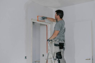 Gri üniformalı ve eldivenli yakışıklı beyaz bir adam, merdiven üzerinde duruyor, bir kapı aralığında süngerimsi bir malayla yeni sıvanmış bir duvarı düzleştiriyor.