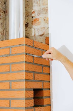 Tanınmayan genç bir beyaz adam, bir eliyle şömine bacasındaki tuğlalar arasında gün boyunca sandalyeye otururken uzun ve dar bir spatula kullanarak taze çimento harcı ile temizlik yapıyor.