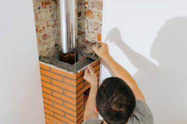 Beyaz tenli genç bir adam taze çimento harcı döşeyen bir mala ve ince bir ahşap bir katman kullanarak şömine bacasında temiz tuğla yapıyor, üst tarafı seçici bir odak ile yakın görüyor. Temiz