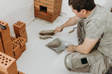 Kafkasyalı genç bir erkek müteahhit, bir süpürgeyle tuğla duvarının ortasında kurumuş çimento kalıntılarını süpürüyor, odada çömelmiş vaziyette faraş gibi tırmıklıyor. kenar görünümü kapat