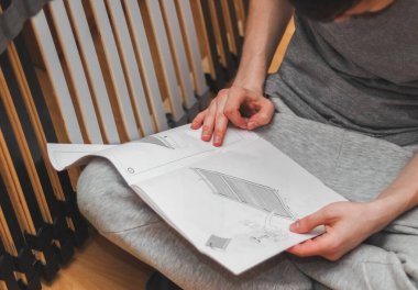 Genç, beyaz bir adam yerde oturmuş, yatak yapmak için kâğıt talimatlarını okuyor. Yan taraftan, saha derinliği ile yakın plan. DIY mobilya montaj konsepti, okuma talimatları.