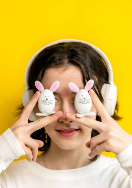 Portrait of one young Caucasian beautiful happy teenage girl holding with her fingers two Easter decorative bunny at eye level, standing on a yellow background on a spring day in the room, side view clipart