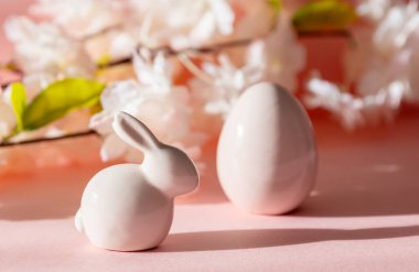 Paskalya yumurtası ve tavşanın porselen heykelcikleri pembe bir arka planda duruyor. Elma ağacı çiçeklerinin bulanık bir dalı, yakın plan görüntüsü var..