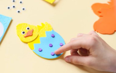 Kafkasyalı bir genç kızın elleri, sarı bir tavuğa, soluk sarı bir tavuğun üzerinde el yapımı bir takım eşyalar bulunan el yapımı bir masada oturan, eflatun rengi küçük bir çember yapıştırıyor.