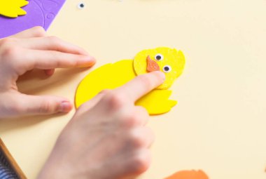 Kafkasyalı genç bir kızın elleri sarı keçeli tavuğun üzerine turuncu bir gaga çıkartması yapıştırıyor parmaklarıyla, soluk sarı arka planda bir dizi el işi ile bir çocuk masasında oturuyor.