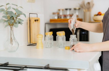 One young Caucasian unrecognizable girl pours seasoning spice black peppercorns from a plastic bag into a new glass transparent jar, standing at a white table in the kitchen on a summer day, side view