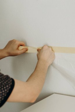 One young unrecognizable Caucasian guy glues with both hands adhesive tape with transparent cellophane on the wall to collect dust and debris, before drilling with a drill, side view close-up.