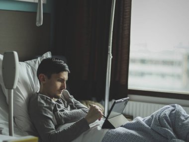 Pencerenin yakınındaki bir hastane odasında tabletle yatan üzgün, beyaz bir gencin portresi..