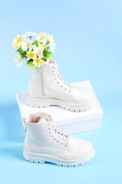 White demi-season martens boots made of eco-leather with a rough sole with a bouquet of spring flowers and a white cardboard box for shoes stand on a light blue background, close-up side view. The clipart