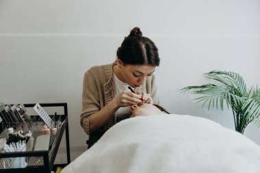 Bir kadın müşterinin sol gözündeki iki cımbızla yapay kirpikleri yapıştıran güzel, beyaz bir kız kozmetik uzmanının portresi.