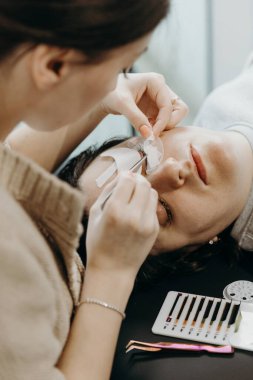 Beyaz tenli güzel bir kozmetik uzmanı kızın portresi. Kozmetolojide yatan bir kadın müşterinin sol gözkapağının alt kısmındaki iki cımbızla kağıt etiketlerini soyuyor.