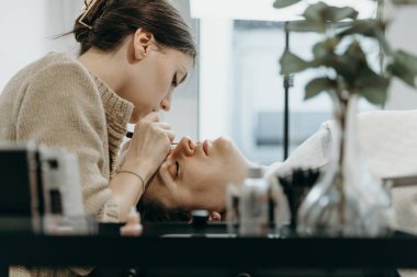 Beyaz tenli güzel bir kozmetik uzmanı kızın portresi. Yapay kirpiklerini, bir kadın müşterinin sol gözündeki iki cımbızla yapıştırıyor. Ev ofisinin kozmetoloji masasında yatıyor.