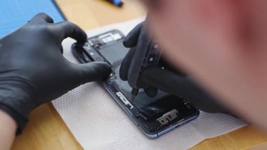 Siyah lateks eldivenli genç bir beyaz adam bir yaz günü mutfakta masada otururken elektrikli tornavida kullanarak akıllı telefonun arkasına küçük bir cıvata vidalıyor.