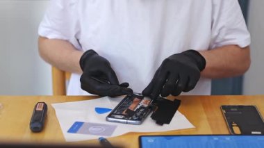 Siyah lateks eldivenli genç bir beyaz adam, bir yaz günü mutfak masasında otururken özel keskin bir tornavida kullanarak akıllı telefonun küçük parçalarıyla bir tabak ayırıyor.