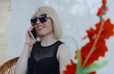 Siyah bluzlu ve güneş gözlüklü genç güzel bir kızın portresi gülümseyen bir akıllı telefonla konuşuyor, hasır bir sandalyede beyaz bir perdenin arkasında oturuyor ve kırmızı bir bulanıklık