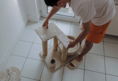 Genç yakışıklı beyaz bir adam elle ikinci kattaki bir rafa vidalıyor kedi evinin karton bir tüpüne, tadilattan sonra, bir yaz günü kapısının yanındaki fayanslı zeminde duruyor.