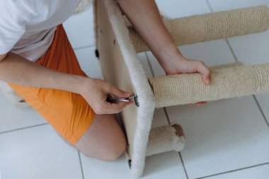 Genç, yakışıklı, tanınmayan beyaz bir adam bir yaz günü fayans zemininde otururken tamir etmek için kedi evinin karton bir tüpünden vidayı elleriyle söker.