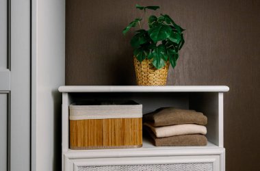 Doğal keçi kılından yapılmış çıplak gözlüklü ince süveterler eski beyaz bir dolabın rafında katlanmış bir şekilde duruyor. Bambu sepetinin yanında. Kahverengi bir duvarın karşısında saksıda çiçek var.