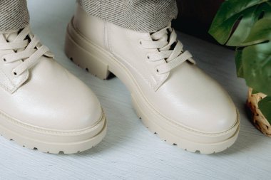 Bir çift şık şık kadın klasik krem rengi botlar kaba tabanlı ve dantelli, hafif beton zeminde yünlü pantolonlar ve duvarın yanında bir çiçek, üst tarafı çok yakın.