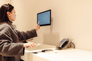 Gri montlu, beyaz tenli genç bir kız, cep telefonuyla KHK kodunu bağımsız olarak tarıyor ve bir bilgisayar ekranını işaret ediyor. Alışveriş merkezindeki bir mağazada duruyor.