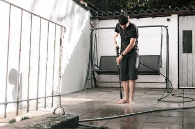 Bir bahar günü gün batımında, evin arka bahçesinde bir kercher kullanarak, basınç altında bahçe fayansları yıkayan esmer, beyaz bir adamın tam boy portresi.