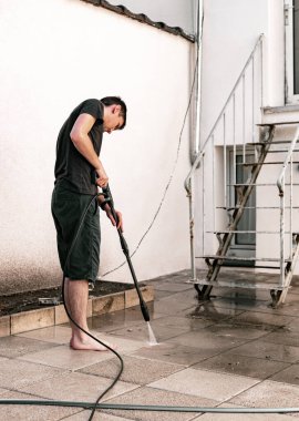 Genç, beyaz bir adamın portresi. Kirli bahçe fayanslarını jetle yıkıyor. Basınçlı çamaşır makinesi kullanıyor.