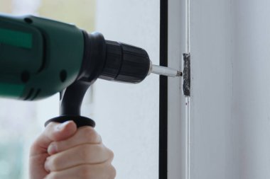 Genç beyaz bir adamın portresi, pencere çerçevesinin metal bir kısmındaki vidayı söken yeşil bir matkapla, alt tarafı çok yakından onaran. Konsept: