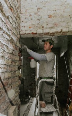 Gündelik giysiler içindeki yakışıklı beyaz bir adam merdiven üzerinde durur ve eski bir evin tavanındaki bir kapıdan tuğla çıkarır. Sprey şişesinden suyla nemlendirir.