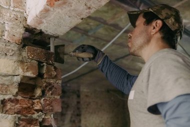 Genç bir beyaz adam tuğlalara spatulayla taze çimento serpiştiriyor, onu düzleştiriyor ve soldaki eski bir evin merdiveni üzerinde duruyor..