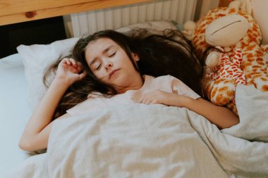 Kafkas esmer, güzel bir kız, tahta bir yatakta uyur. Yumuşak oyuncak bir zürafa, radyatörün yanında gri bir battaniyeyle kaplıdır..