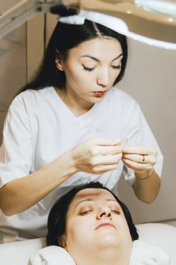 Beyaz önlüklü bir kozmetik uzmanı olan beyaz tenli güzel bir esmer kızın portresi boyama ve kirpikleri kaplama işlemine başlamadan önce bir kağıt çıkartmasını soyuyor.