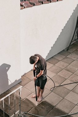 Beyaz adamın tam boy portresi. Kirli bahçe fayanslarını basınçlı su jeti ile yıkıyor. Basınçlı çamaşır makinesiyle.