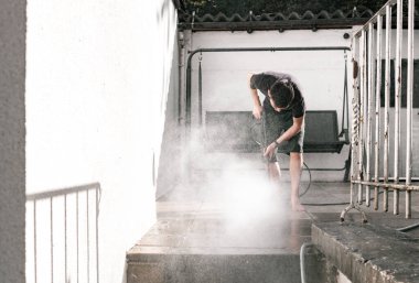 Genç beyaz bir adamın portresi bahçe fayanslarını basınç makinesinden çıkan bir su püskürtüsüyle yıkıyor, küçük sıçramalara uçuyor, bir bahar günü arka bahçede eğimli bir pozisyonda duruyor,