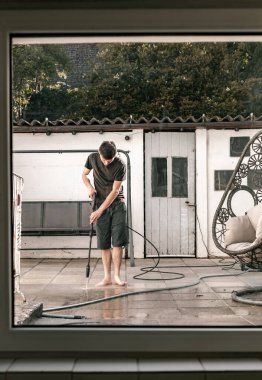 Koyu renk elbiseli yakışıklı beyaz adamın tam boy portresi. Yalınayak duruyor ve bahçedeki kirli fayansları temizliyor. Basınçlı su jeti ile bahar günü kercher kullanıyor.
