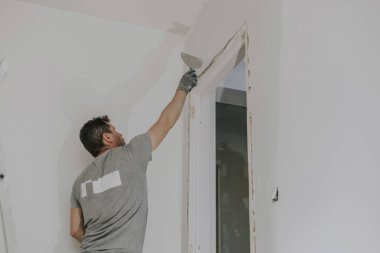 Gri üniformalı ve eldivenli yakışıklı beyaz bir adam, merdivenlerde dikilirken çürümüş bir kapı eşiğinde küçük bir spatulayla çöpleri fırçalıyor. İnşaat manzarası aşağıda.
