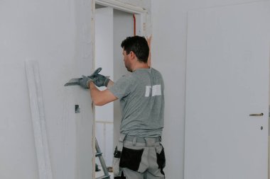 Gri üniformalı genç beyaz bir adam, bir tamir odasının kapısında dikilirken, büyük bir malayla, beyaz bir macunla, seçici bir odak noktasıyla, yan görünüme yakın bir şekilde yaklaşır. Şey...