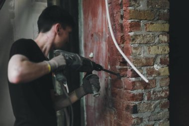 Siyah üniformalı, genç, beyaz bir adam. Tuğla duvarda bir delik açıyor. Büyük bir tornavidayla deliniyor. Ev kavramı