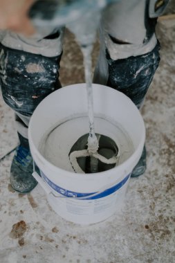 Beyaz tenli, tanınmaz halde beyaz macunu beyaz bir kovada, küçük bir inşaat mikseriyle kaplayan, kirli bir evin içinde duran, üst tarafı seçici bir odak ile yakın çekim yapan genç bir erkek. Konsept