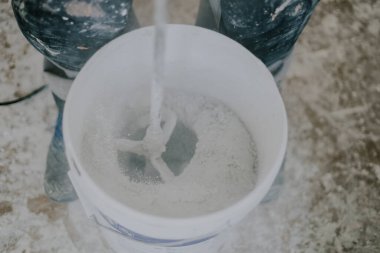 Young caucasian male unrecognizable mixing white dry putty with a wet construction mixer in a white small bucket with flying dust, standing on a dirty floor indoors, close-up top view with selective