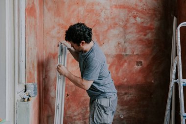 Kıvırcık saçlı, yakışıklı beyaz bir adam plastik bir pencere çerçevesinin üzerine kağıt peçete silerken tamiratın devam ettiği bir odada yan yana duruyor.