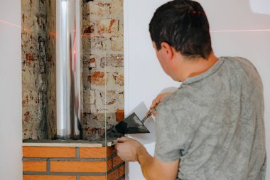 Genç bir beyaz adam şöminenin bacasındaki bir tuğlaya beton havan topuyla beton harcı uyguluyor ve onu tahta bir batten ile düzleştiriyor. Gün boyunca odada duruyor.