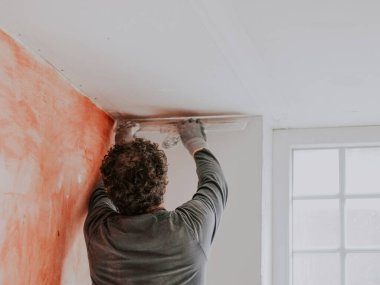 Üniformalı, kıvırcık saçlı, genç bir beyaz adam, pencerenin yakınındaki duvardan macunları çekiyor. Büyük bir spatulası var. Merdiven üzerinde duruyor.