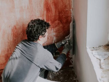 Gri tişörtlü ve kıvırcık kahverengi saçlı genç beyaz bir adam eşitlik için kontrol ediyor. Pencerenin altındaki duvarda elinde büyük bir spatula olan yeni bir macun var. Şey...