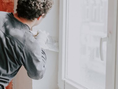 A caucasian young guy a plasterer in a gray T-shirt with sleeves and curly brown hair levels fresh putty with a spatula on a window opening, side view close-up. The concept of repairing apartments and