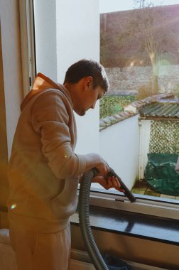 Genç beyaz bir adamın diz boyu portresi. İkinci kattaki pencere çerçevelerinden enkazı vakumluyor. Güneşli bir günde pencereyi temizlemeye hazırlanıyor. Konsept