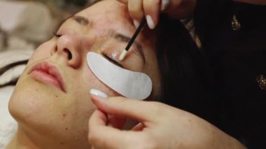 Genç bir müşteri için pamuk çubuğu ve sıvı çözeltisi kullanarak sol göz kapaklarını dikkatlice temizleyen beyaz bir kozmetikçi kız bir güzellik salonunda yatıyor.