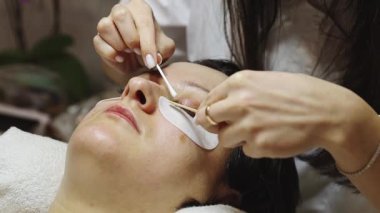 Kalıbı bitirdikten sonra sol gözünün kirpiklerini pamukla ve tahta sopayla tarayan genç bir beyaz kız kozmetik uzmanı. Bir müşterisi güzellik salonunda kozmetoloji masasında yatıyor.