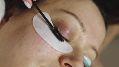 Sağ gözünün kirpiklerini siyah bir fırçayla fırçalayan beyaz bir kız kozmetik uzmanının elleri. Bir müşterisi, bir güzellik salonunda kozmetoloji masasında yatıyor.