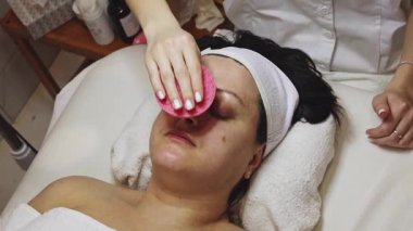Genç bir doktor kozmetik uzmanı, bir müşterisinin alnını ve çenesini pembe bir süngerle temizler. Pembe süngerle. Güzellik salonundaki masaj masasında gözleri kapalı yatar.